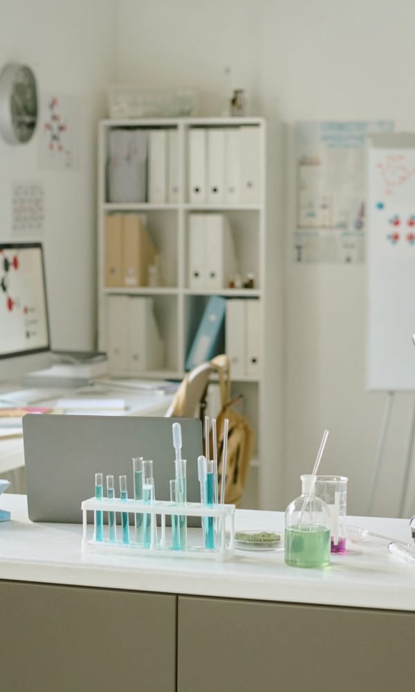 Well-lit science laboratory with computers, glassware, microscopes, and books neatly organized on tables and shelves surrounding the workspace