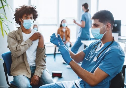 Coronavirus Immunization. Doctor With Syringe Taking Dose Of flu Vaccine For Injection From Vial, Holding Bottle Wearing Blue Latex Gloves And Protective Mask, Patient Waiting In Background