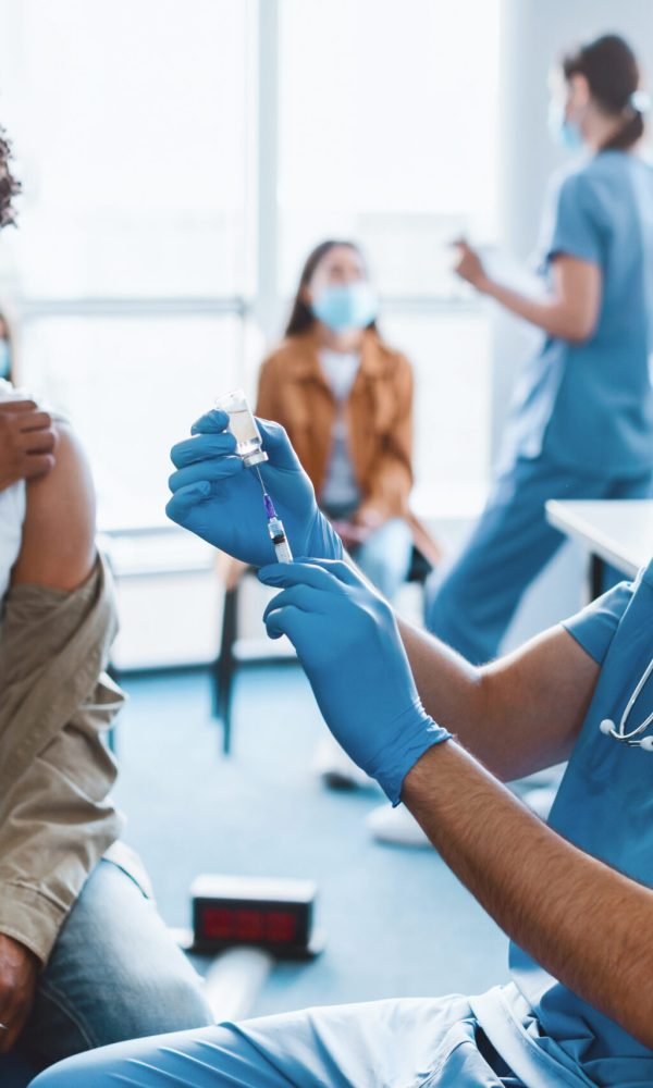 Coronavirus Immunization. Doctor With Syringe Taking Dose Of flu Vaccine For Injection From Vial, Holding Bottle Wearing Blue Latex Gloves And Protective Mask, Patient Waiting In Background
