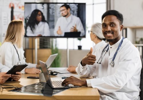 African american male physician with stethoscope working on laptop during online conference in hospital. Bearded man looking at camera while listening presentation with team of doctors on background